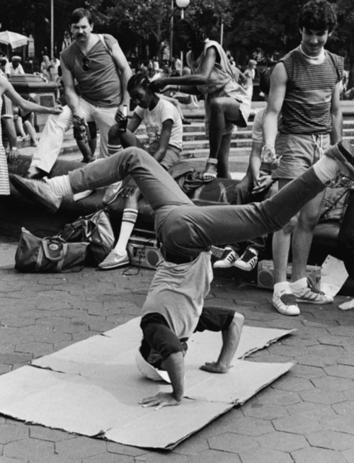 Black-and-white photo of a breakdancer spinning on a cardboard mat in a city park while a crowd watches.